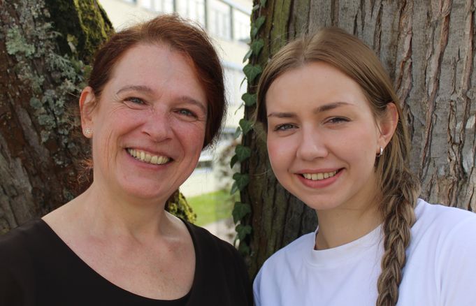 Zwei lächelnde Frauen schauen in die Kamera vor Baumstämmen